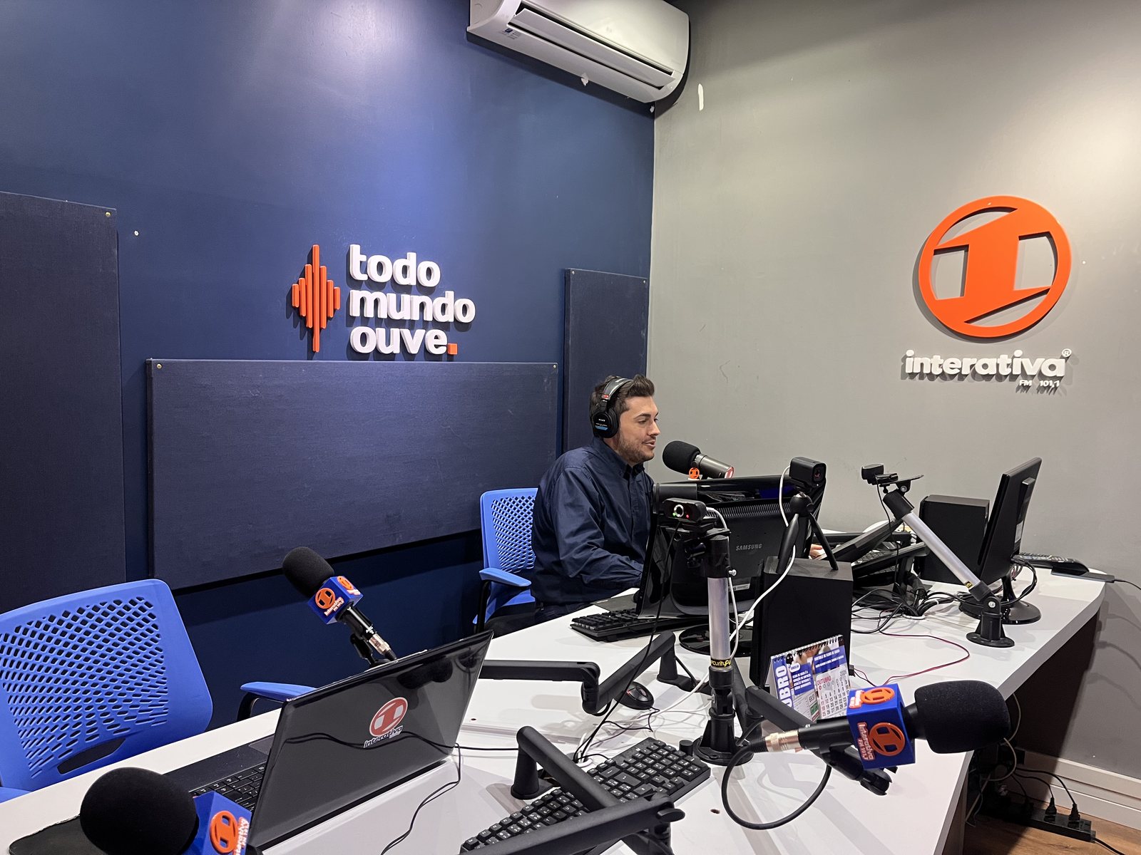 Locutor no ar no estúdio da Rádio Interativa FM, com a marca e o slogan todo mundo ouve na parede