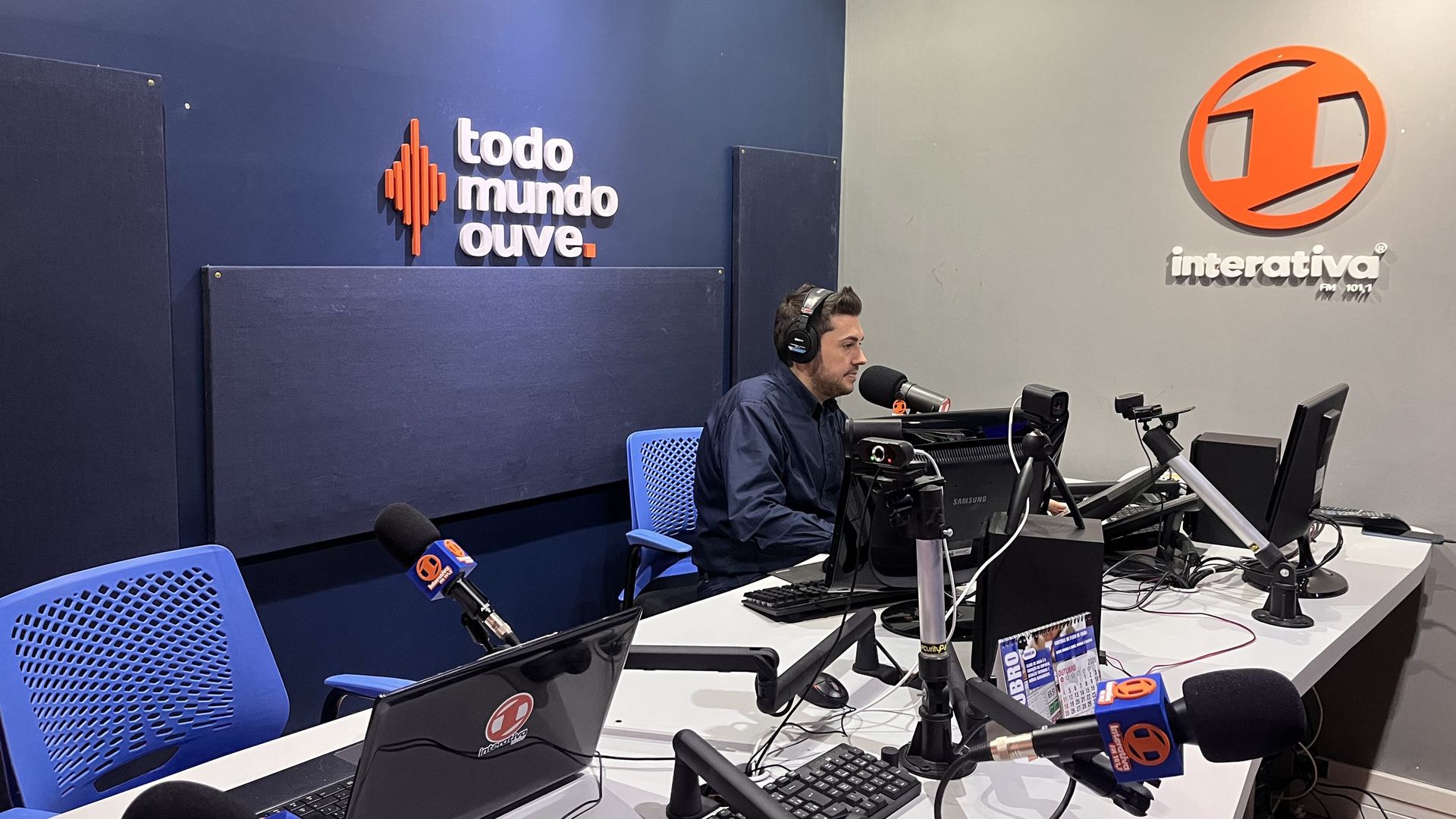 Locutor no ar no estúdio da Interativa FM, com a marca e o slogan todo mundo ouve na parede