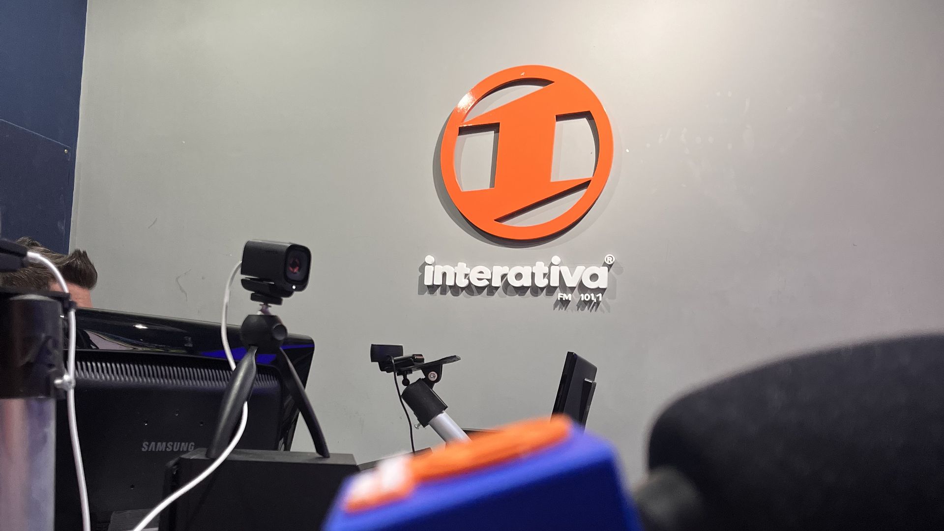 Microfone com a marca da Interativa FM em primeiro plano e o logo da rádio na parede do estúdio