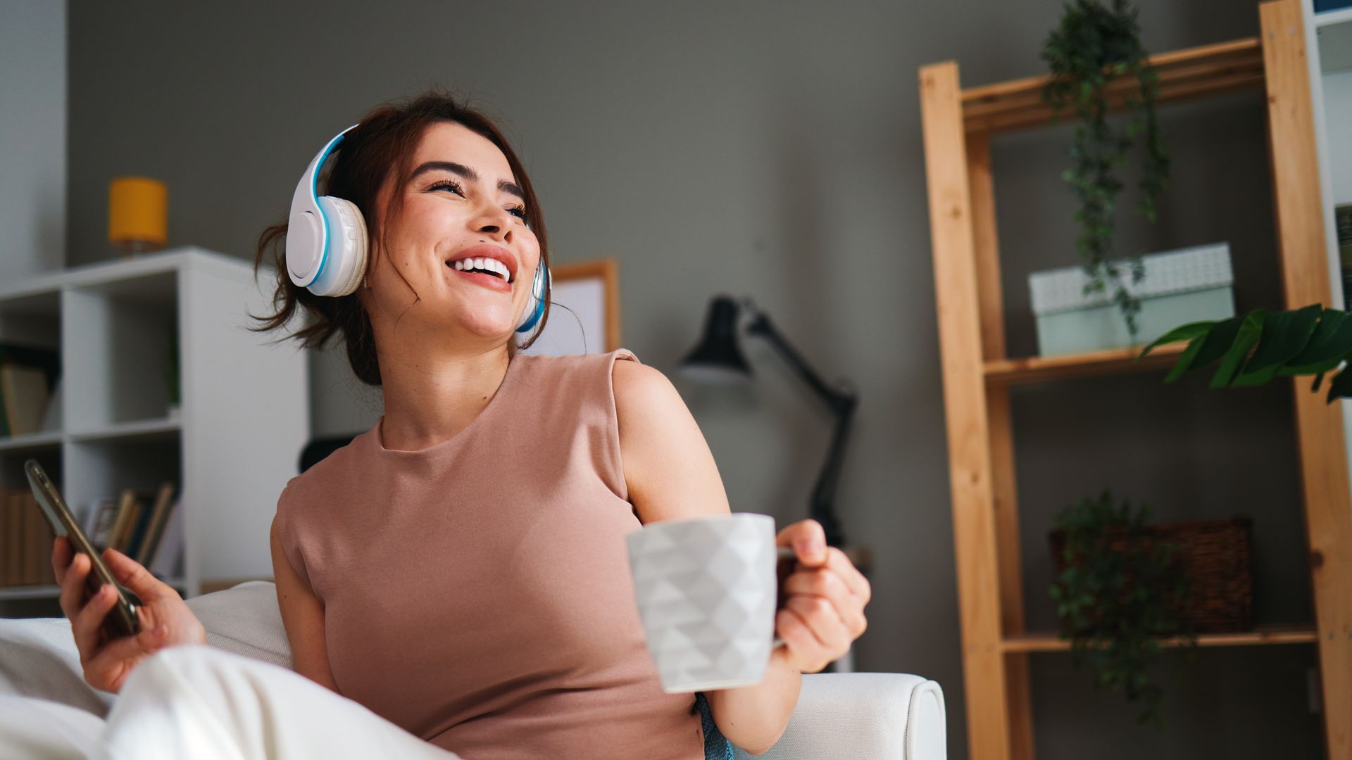 Mulher sorrindo, de fone de ouvido, ouvindo rádio em casa com uma xícara na mão